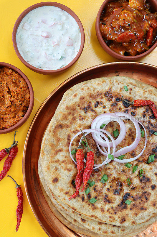 金属托盘上放着aloo paratha，上面撒着辣椒、红洋葱圈和切碎的韭菜，美味的面饼里塞满了捣碎的garam masala土豆、酸辣酱、泡菜蘸酱和raita(酸奶)，俯瞰美景